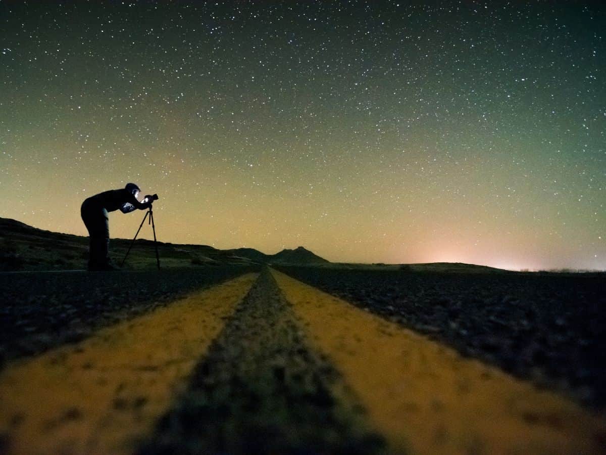 A man is standing on a road with a camera under a starry sky.