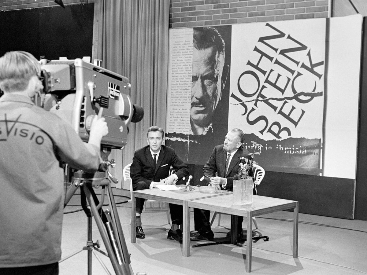 A black and white photo of writer John Steinbeck being interviewed in a Helsinki studio.