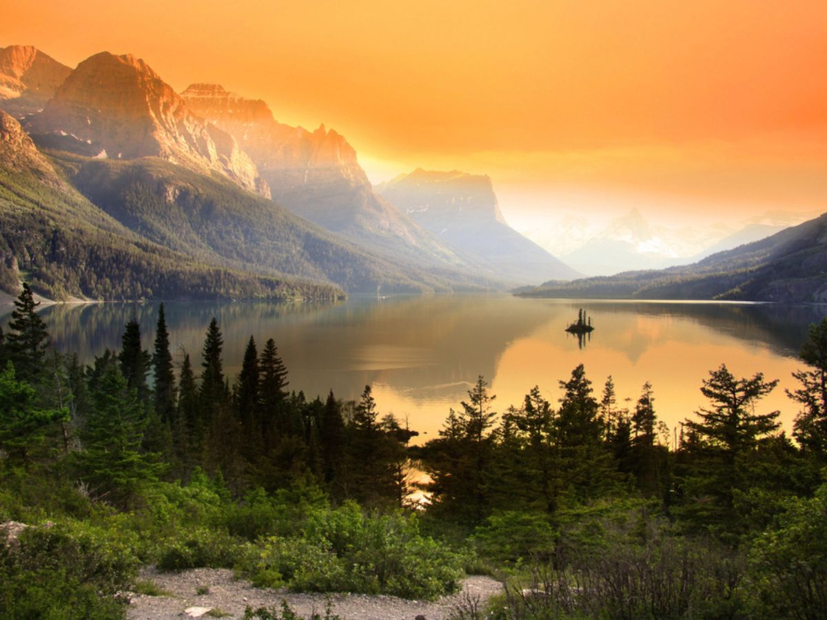 Sunset over a serene mountain lake with a forest in the foreground.