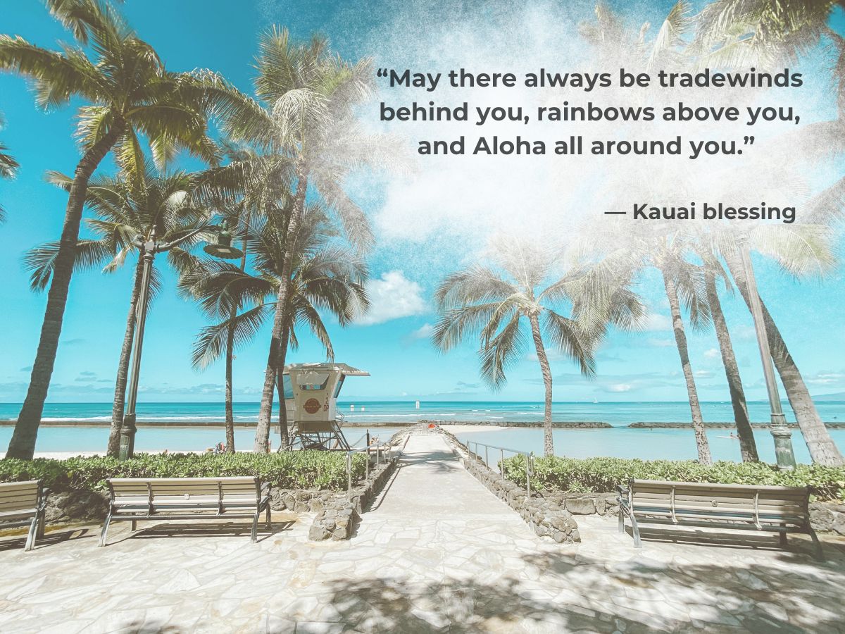 Walkway leading to a beach with palm trees and a lifeguard tower under a clear sky, accompanied by a kauai blessing quote.
