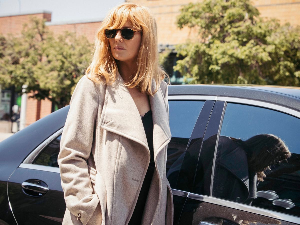 Beth Dutton in sunglasses and a beige coat stands by a black car, her expression serious and confident.