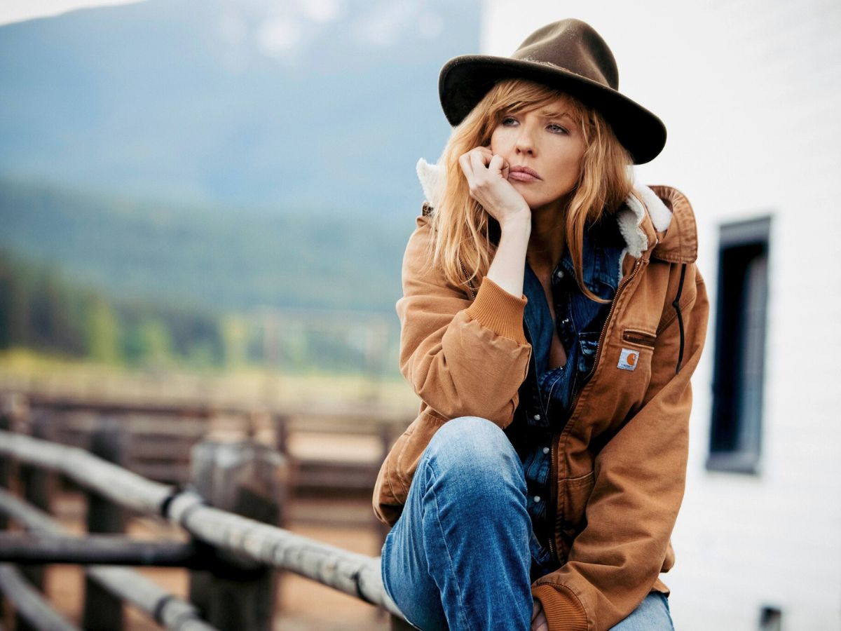 Beth Dutton in a brown hat and denim jacket sitting on a wooden fence with mountains in the background.