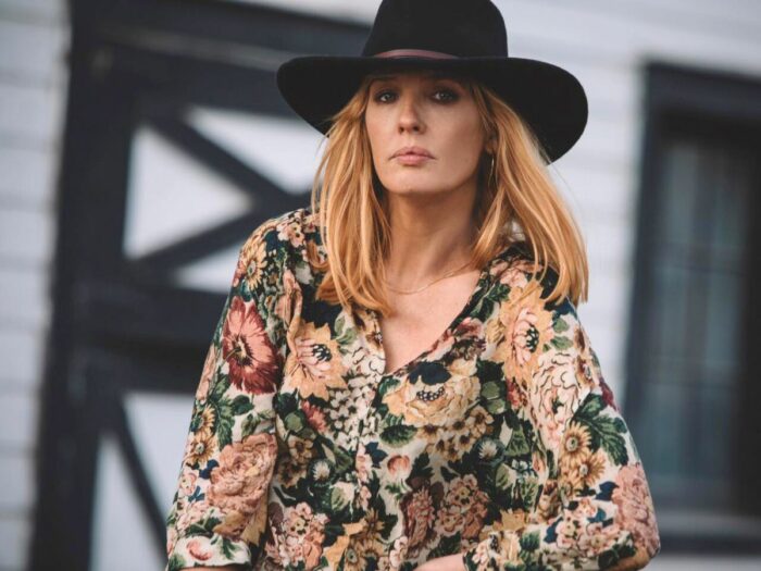 Beth Dutton wearing a floral blouse and a black wide-brimmed hat stands confidently, with a focused expression, in front of a barn.