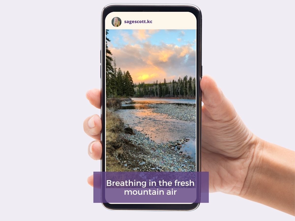 Hand holding a smartphone displaying a mountain river landscape at sunset with the caption "breathing in the fresh mountain air.