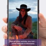 A person holding a smartphone displaying an instagram post with a photo of a woman in a hat sitting outdoors by a campfire, featuring a caption about wandering in montana.