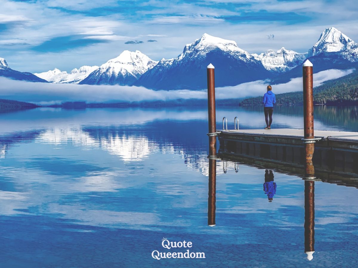 A person standing at the end of a dock overlooking a calm lake with mountains in the background.